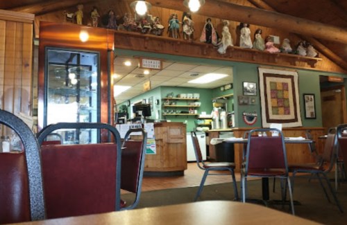Cozy restaurant interior with wooden decor, tables, and shelves displaying dolls and a quilt on the wall.