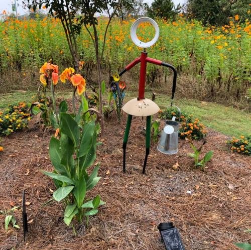 A whimsical garden figure made of metal, holding a watering can, surrounded by colorful flowers and greenery.