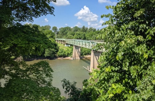 A green bridge spans a river, surrounded by lush trees and a clear blue sky with fluffy clouds.