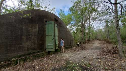 A person stands beside a large, weathered green door on a concrete structure, surrounded by trees and a gravel path.