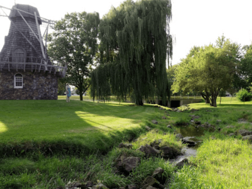 A scenic view featuring a windmill, lush green grass, trees, and a small stream in a tranquil landscape.