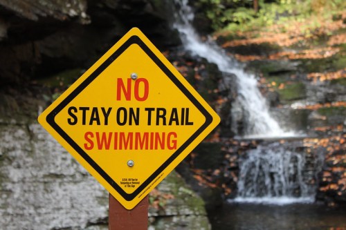 A yellow warning sign reads "No Swimming" and "Stay on Trail" in front of a waterfall and rocky landscape.