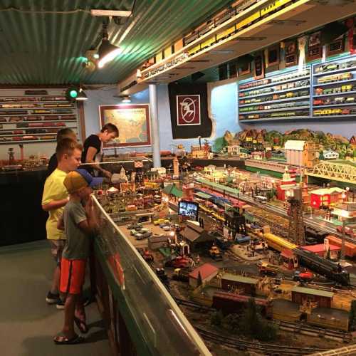 Children observe a detailed model train display with various buildings and landscapes in a well-lit room.