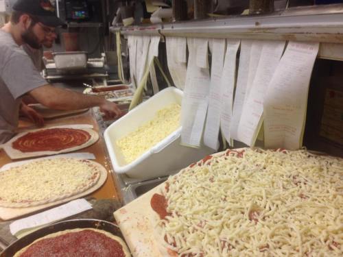 A busy pizza kitchen with a chef preparing pizzas, topped with sauce and cheese, surrounded by order tickets.