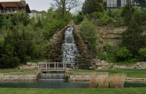 A serene waterfall cascades down a rocky structure, surrounded by lush greenery and a calm pond.