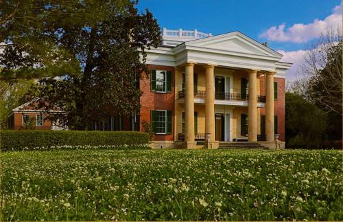 A grand two-story brick mansion with columns, surrounded by a lush green lawn and trees under a blue sky.