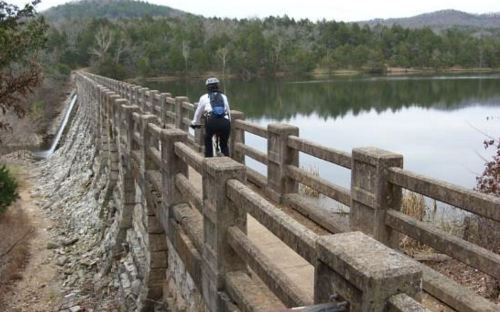 A cyclist on a stone dam overlooking a calm lake, surrounded by trees and hills in the background.