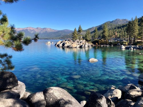 A serene lake surrounded by trees and mountains, with clear blue water and smooth rocks along the shore.