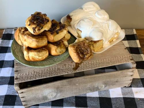 A wooden crate holds a plate of cinnamon rolls with icing and a stack of sticky buns on a checkered tablecloth.