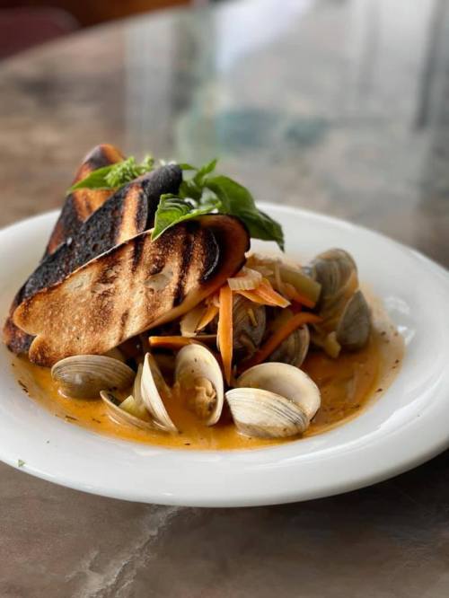 A plate of clams with sautéed vegetables and charred bread, garnished with fresh basil on a marble table.
