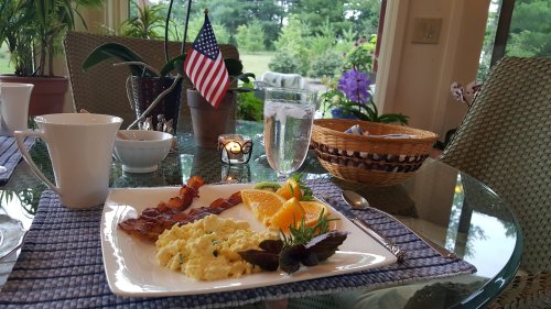 A breakfast plate with scrambled eggs, bacon, fruit, and a glass of water, set on a table with a small American flag.