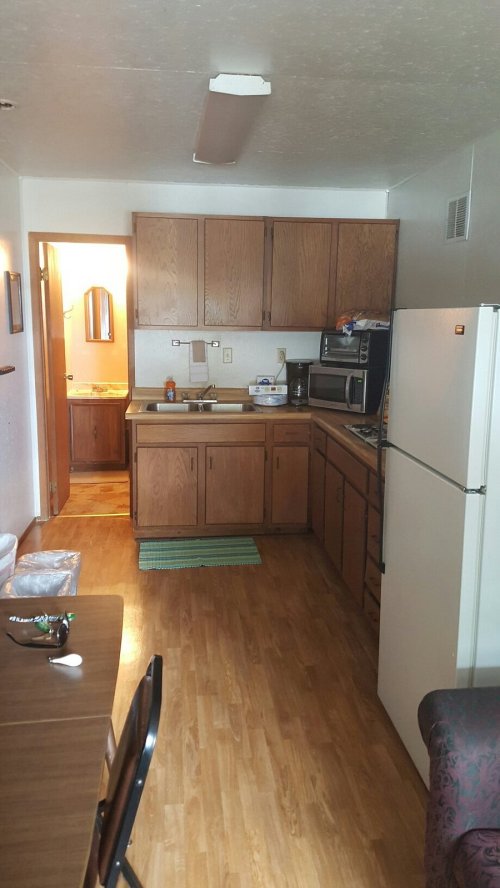 A small kitchen with wooden cabinets, a refrigerator, and a dining table, featuring a doorway to a bathroom.