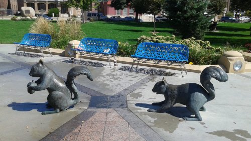 Two bronze squirrel sculptures on a plaza, surrounded by grass and blue benches in a park setting.