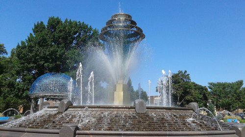 A large, modern fountain with multiple water jets, surrounded by trees and a clear blue sky.