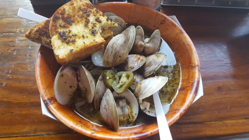 A wooden bowl filled with clams in broth, garnished with jalapeños, and served with toasted bread on the side.