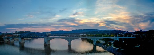 A panoramic view of a bridge over a river at sunset, with colorful clouds and hills in the background.
