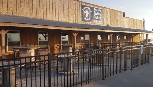 Exterior of a wooden building with a sign for the "Testicle Festival," featuring outdoor seating and a fenced area.