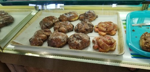 A display of freshly baked pastries, including glazed donuts and apple fritters, on a tray in a bakery.