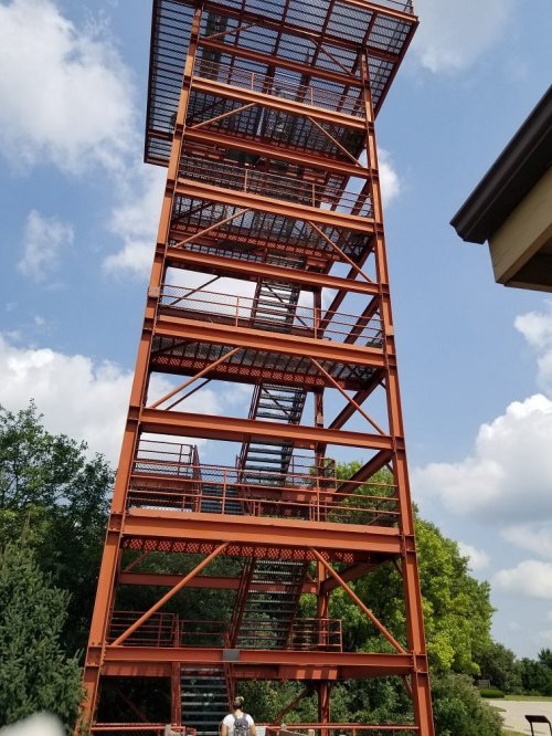 A tall, orange observation tower with stairs, surrounded by greenery and under a partly cloudy sky.