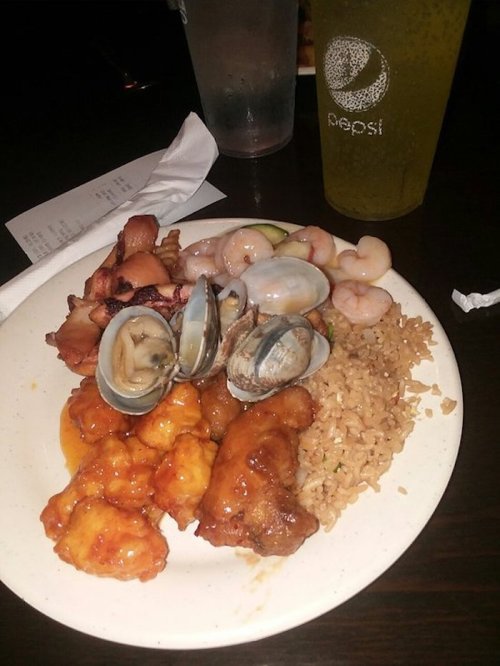 A plate of food featuring fried chicken, seafood, rice, and a drink, set on a dark table.