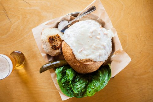 A bowl of clam chowder served in a bread bowl, accompanied by a pickle, lettuce, and a toasted bun on a wooden table.