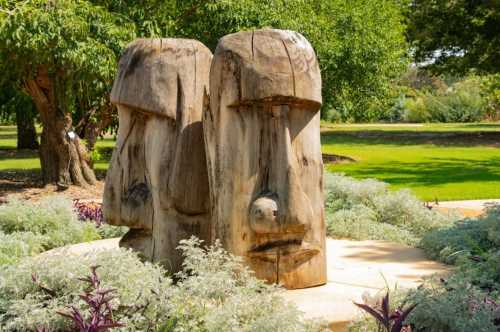 Two large wooden sculptures resembling Easter Island heads, surrounded by greenery and colorful plants in a garden setting.