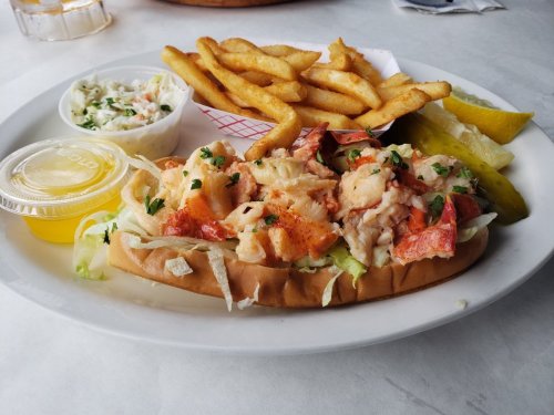 A plate featuring a lobster roll on lettuce, fries, coleslaw, pickles, and a side of butter sauce.
