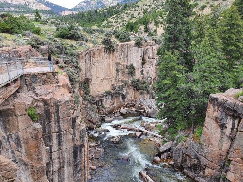 A rocky canyon with a flowing river, surrounded by trees and steep cliffs under a cloudy sky.