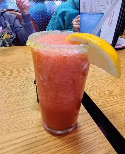A tall glass of frozen pink drink with a sugared rim and a lemon slice on the edge, sitting on a wooden table.