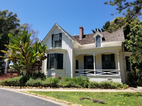 A charming white Victorian-style house with green shutters, surrounded by lush greenery and a clear blue sky.