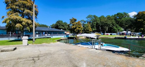 A sunny lakeside scene with a docked boat, grassy area, and a building surrounded by trees.