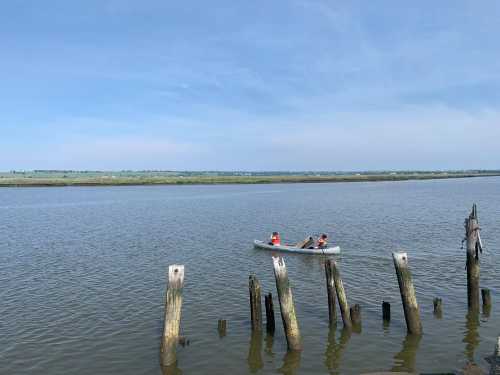 Two people in a small boat on a calm river, with weathered wooden posts in the foreground and green fields in the background.