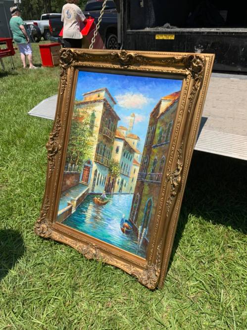 A framed painting of a scenic canal with buildings and a gondola, set on grass near a truck.
