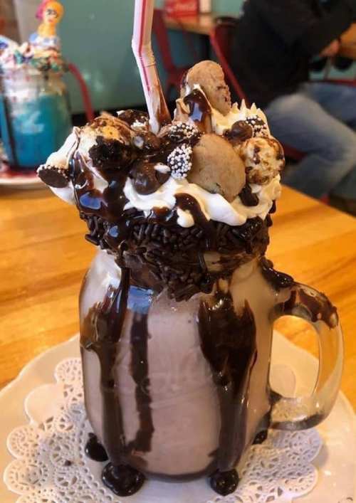 A decadent milkshake topped with whipped cream, chocolate drizzle, cookies, and sprinkles in a glass mug.