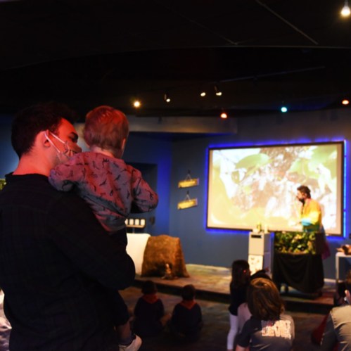 A parent holds a child while watching a presentation in a dimly lit room with a large screen and audience.