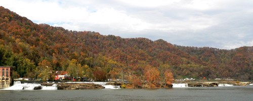 A scenic view of a river with waterfalls, surrounded by autumn-colored trees and rolling hills under a cloudy sky.