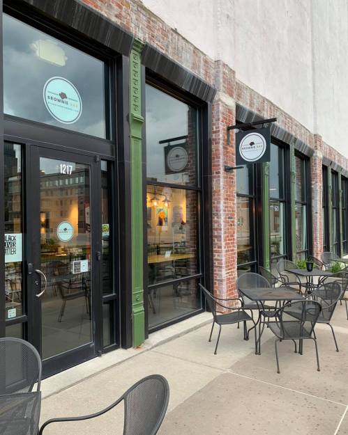 Exterior of a café with large windows, brick walls, and outdoor seating. Signs read "Brownie Bar" and "Black Lives Matter."