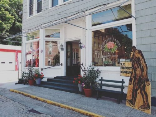 A quaint storefront with large windows, featuring a Bigfoot museum sign and flower pots on the steps.