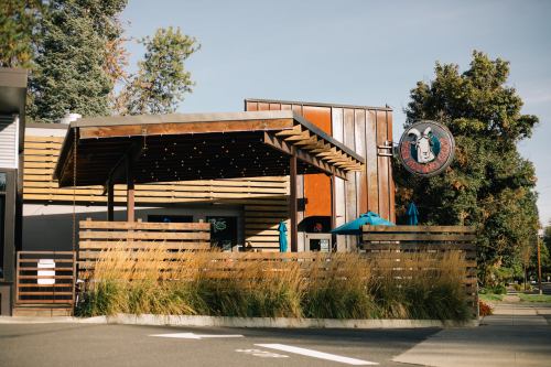A modern restaurant with wooden accents, outdoor seating, and a sign featuring a logo, surrounded by greenery.