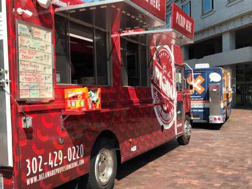 A red food truck with a menu board parked in a plaza, with another food truck visible in the background.