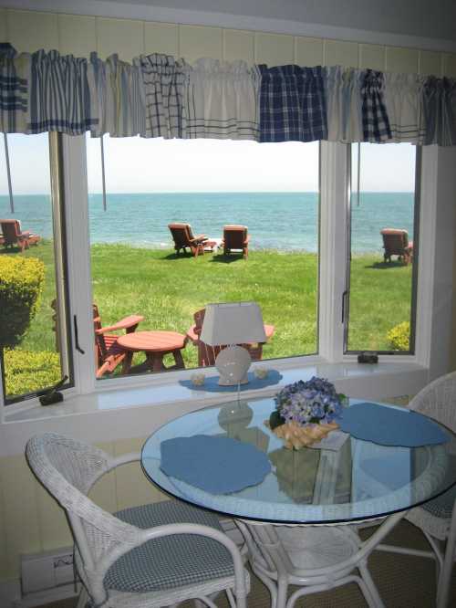 A cozy dining area with a round glass table and wicker chairs, overlooking a serene lake and green lawn.