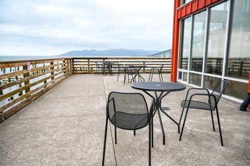 Outdoor seating area with black tables and chairs overlooking a cloudy waterfront and distant mountains.