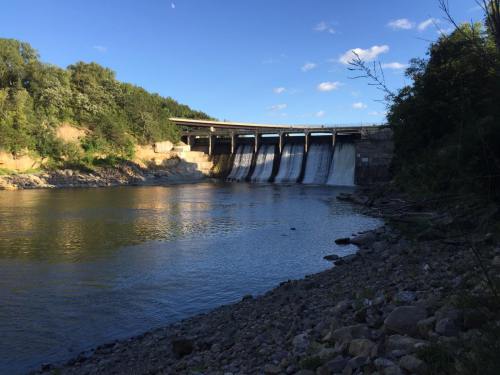 A dam with water cascading over it, surrounded by trees and a calm river under a blue sky with scattered clouds.