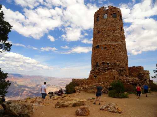 A stone tower stands on a rocky outcrop with visitors exploring the area, set against a backdrop of clouds and canyon views.