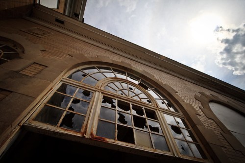 A close-up of a broken window in an old building, with cloudy skies visible through the shattered glass.