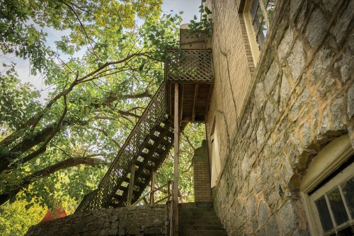 A stone building with a wooden staircase surrounded by lush green trees under a bright sky.