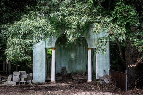 An overgrown structure with two columns, partially hidden by foliage, surrounded by scattered stones and trees.
