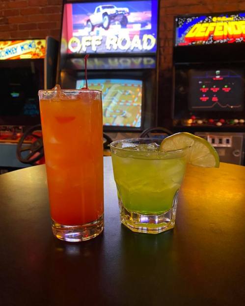 Two colorful cocktails on a table, with a retro arcade game backdrop featuring classic arcade machines.