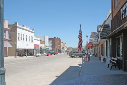 A quiet small-town street lined with shops, an American flag, and clear blue skies.