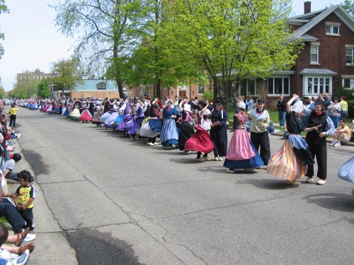 A vibrant parade scene with dancers in colorful skirts performing along a tree-lined street, with spectators watching.